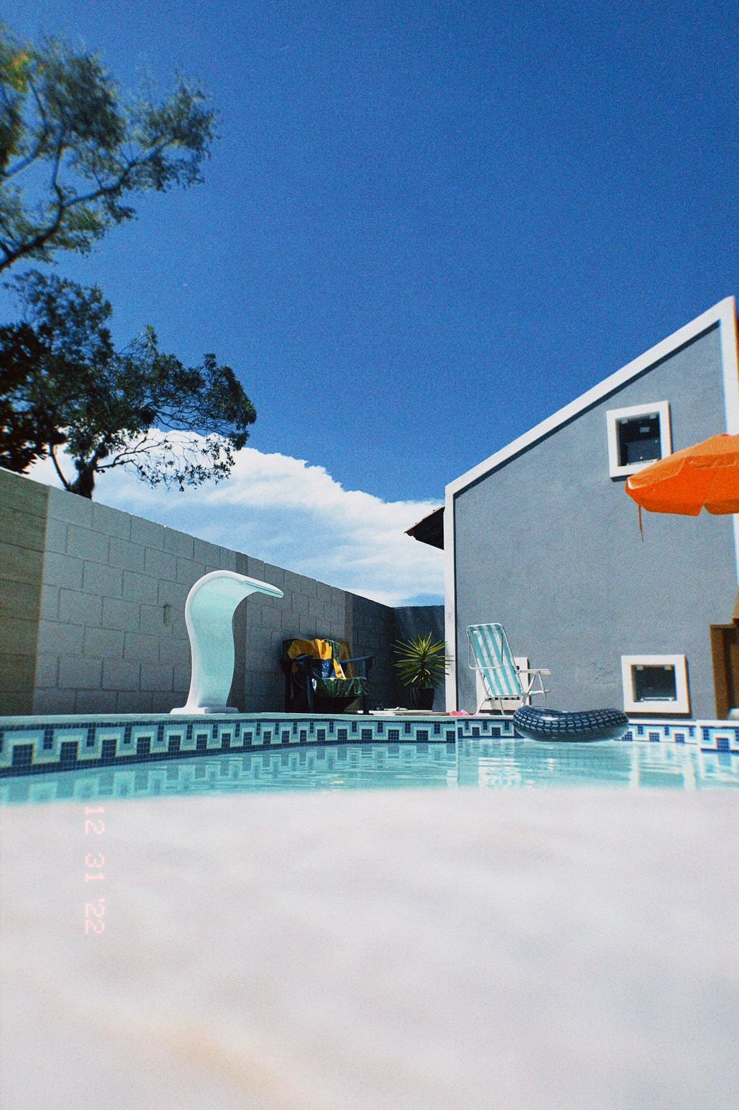 A refreshing swimming pool is shown, surrounded by smooth stone tiles. A vibrant orange umbrella provides shade, while a light blue chair and a decorative element are positioned nearby. The sky is clear, emphasizing the serene outdoor space.
