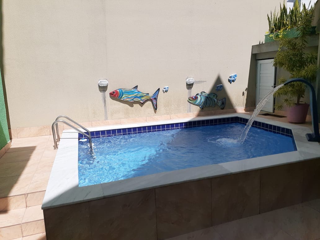 A small, inviting pool is observed, featuring a blue-tiled border and decorative fish murals on a light-colored wall. A gentle water fountain flows into the pool, surrounded by smooth stone flooring and potted plants, creating a serene outdoor space.