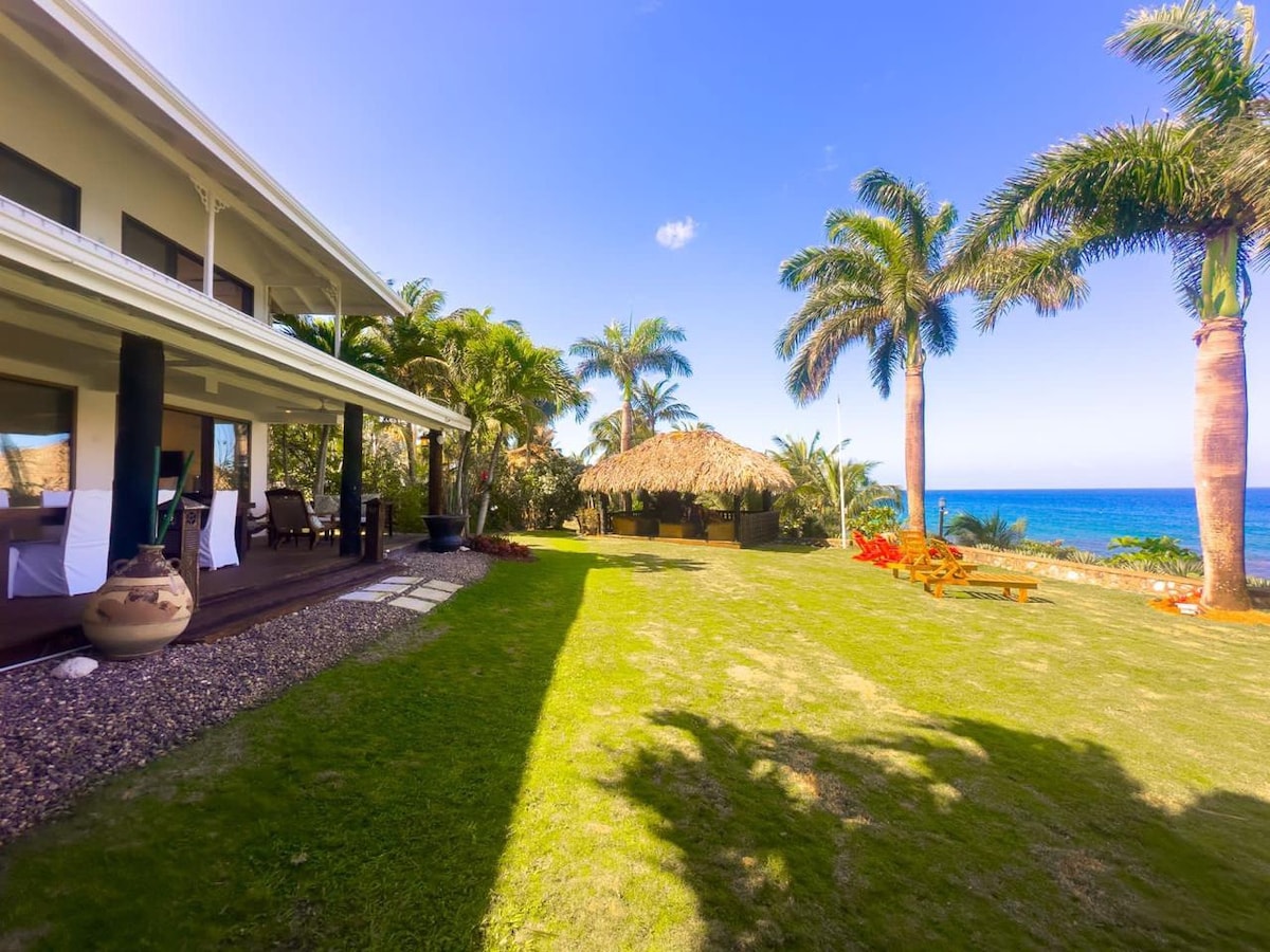 A spacious lawn area is bordered by tall palm trees, leading to a thatched-roof gazebo overlooking the ocean. The villa's outdoor seating area is highlighted, with comfortable lounge chairs positioned to enjoy the scenic coastal views.