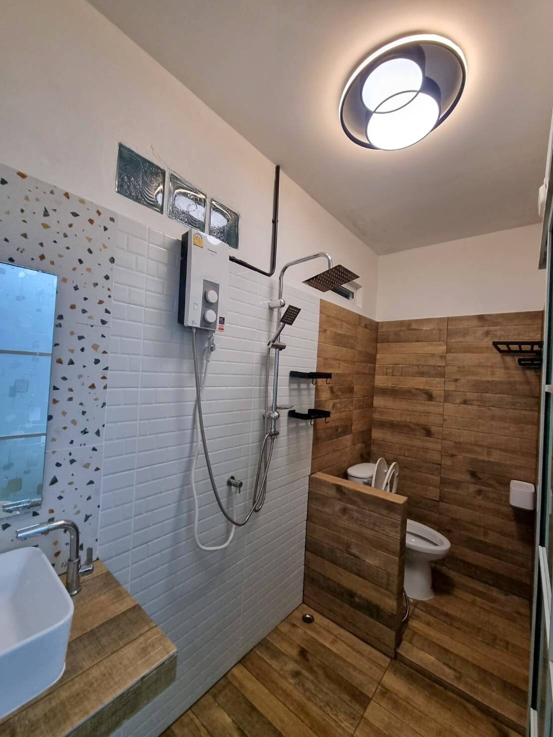 A modern bathroom features a shower area with both handheld and overhead showerheads. A sleek sink sits against a tiled wall, complemented by wooden accents throughout. The space is well-lit by a ceiling light and includes a compact toilet and storage shelves.