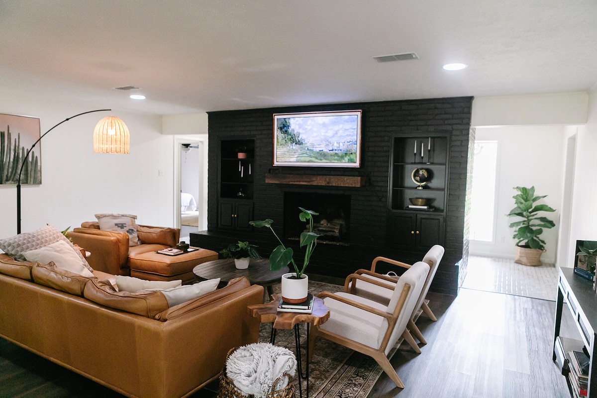 A spacious living area is highlighted by a large, wall-mounted television above a wooden mantel. Comfortable seating includes a leather couch and two armchairs, complemented by a side table with a potted plant. Soft lighting enhances the cozy ambiance of the room.