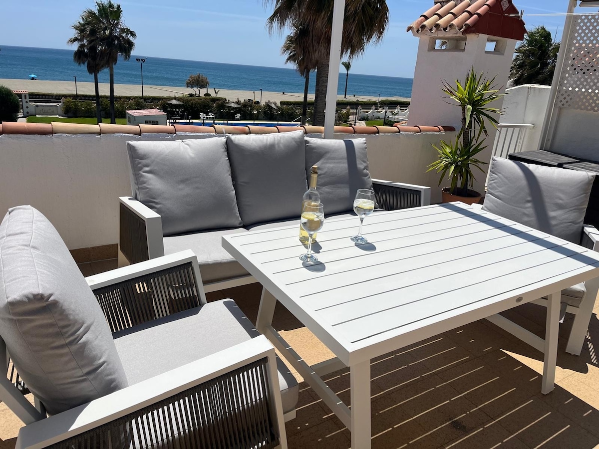 An outdoor terrace is presented with a white table and four cushioned gray chairs. A bottle and two glasses are placed on the table. The view features a pristine beach and palm trees, with the ocean visible in the background.