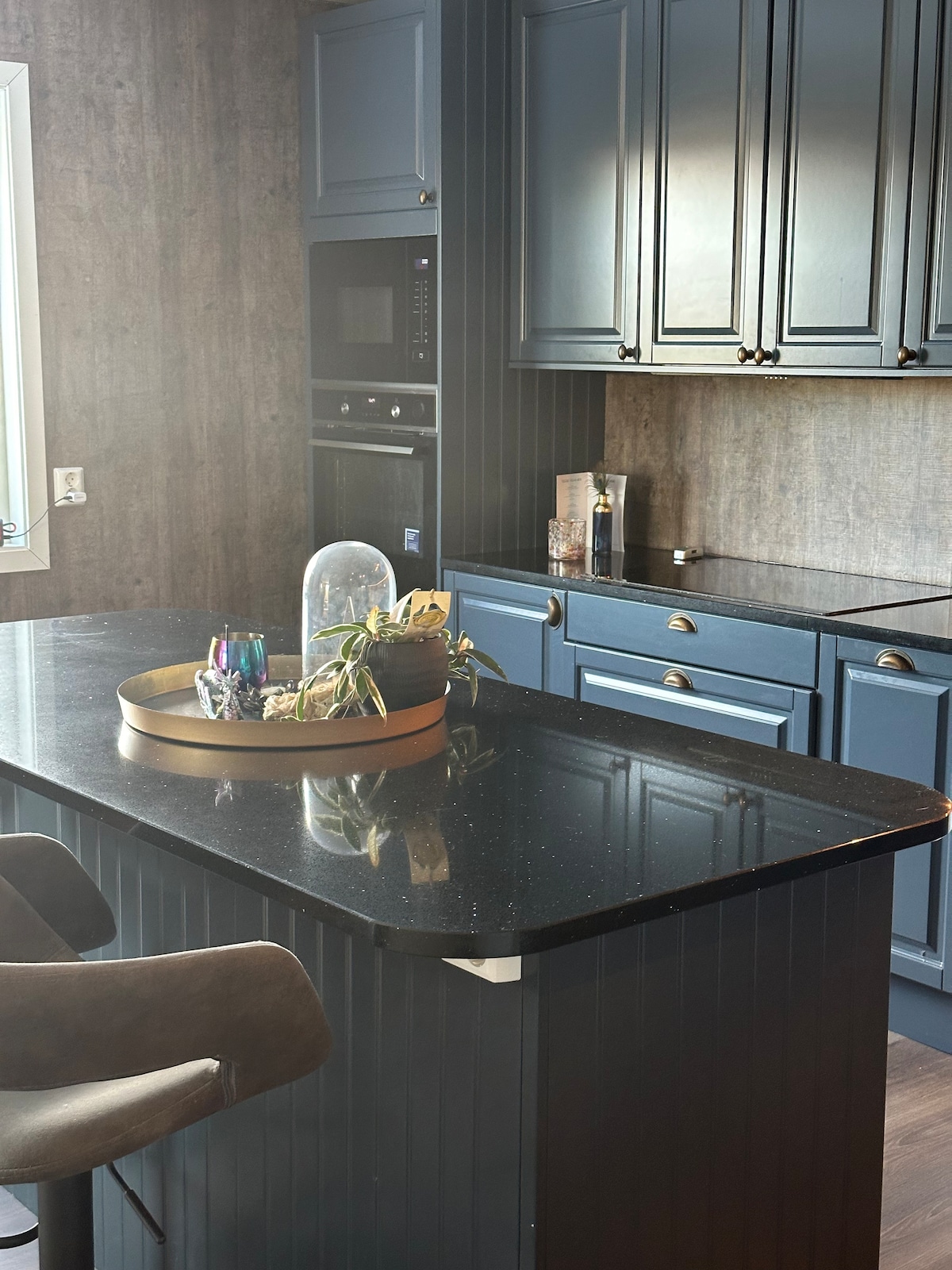 A modern kitchen features dark cabinetry and a spacious island with a polished black countertop. Natural light streams through a window, illuminating decorative plants and a tray of assorted items. Ample storage is provided by upper and lower cabinets.