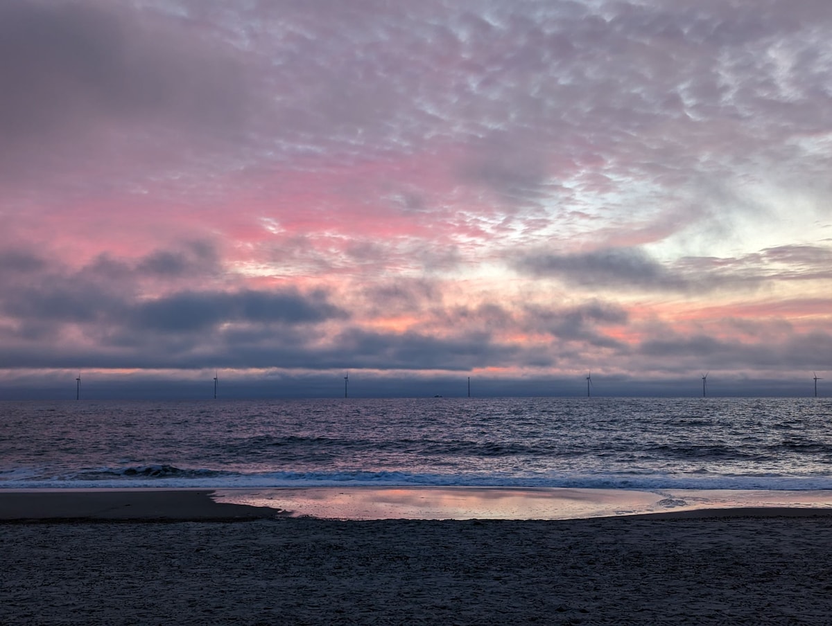 A serene coastal view features a tranquil sea reflecting soft hues of pink and purple in the sky. Dark clouds gently blanket the horizon, with wind turbines visible in the distance. A small pool of water rests on the sandy beach, inviting contemplation.