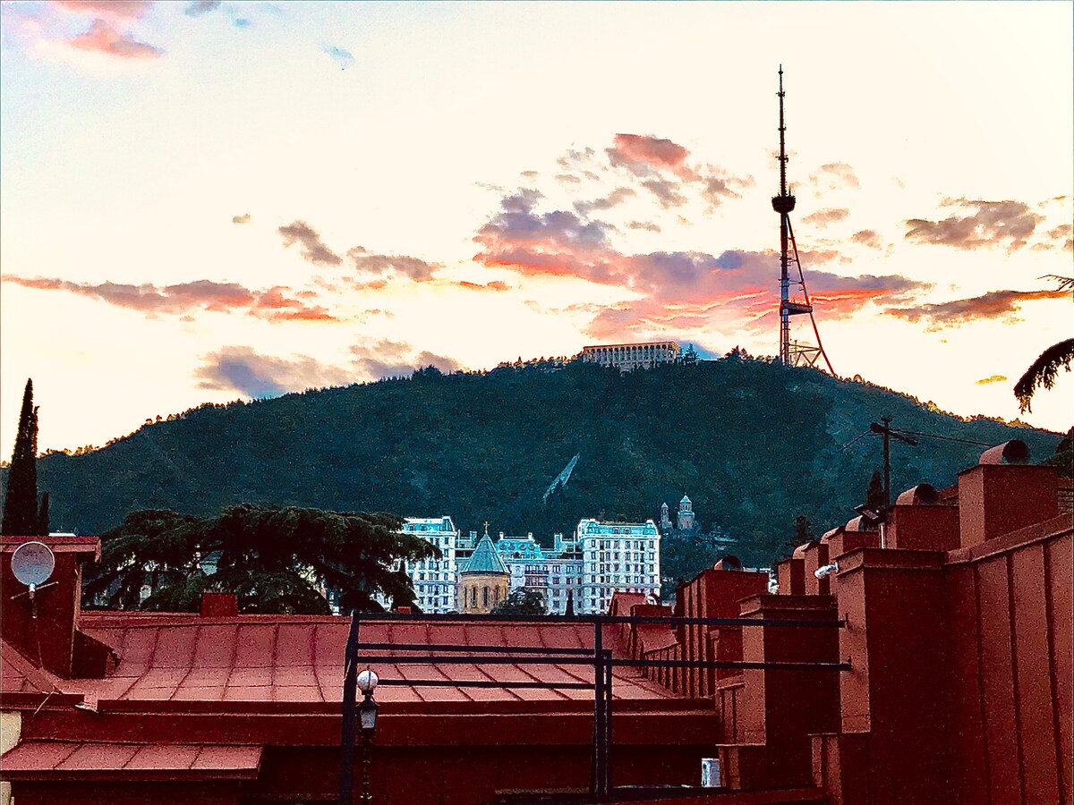 A scenic view showcases a mountain silhouette against the backdrop of a colorful sky at sunset. The green hillside is complemented by the prominent television tower, with rooftops of nearby buildings visible in the foreground.