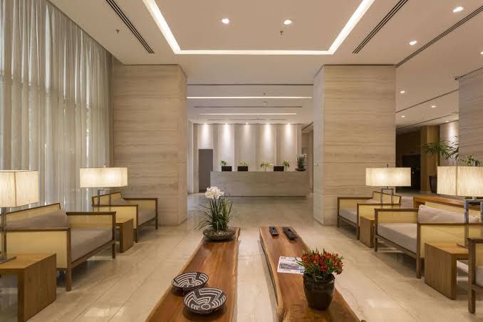 A spacious hotel lobby is presented with light-colored marble flooring and warm wooden accents. Comfortable seating areas with contemporary designs are complemented by minimal decorative elements, including plants and ambient lighting that creates a serene environment. The front desk is positioned prominently in the background.