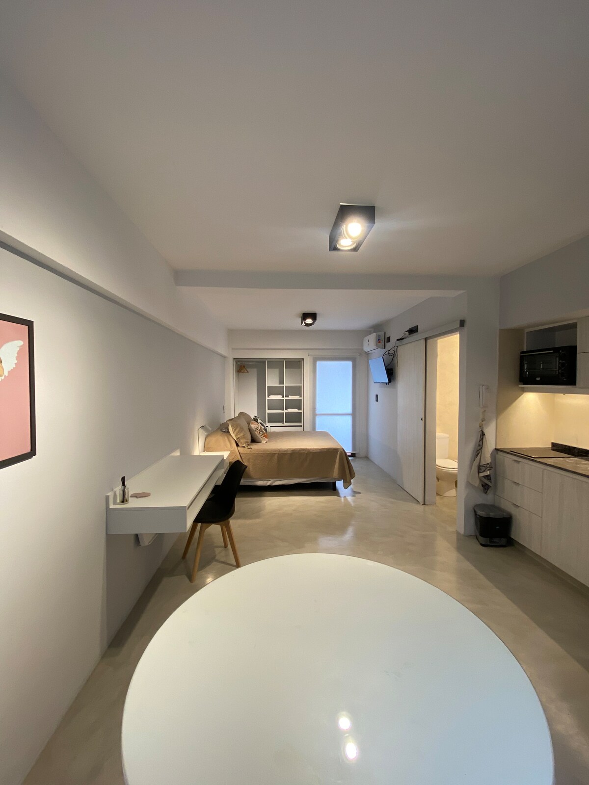 A modern space features a queen-sized bed dressed in neutral tones, with a small table and chair positioned nearby. The kitchenette is equipped with cabinets and appliances, while the bathroom is partially visible. Soft lighting enhances the open layout that combines living and sleeping areas.