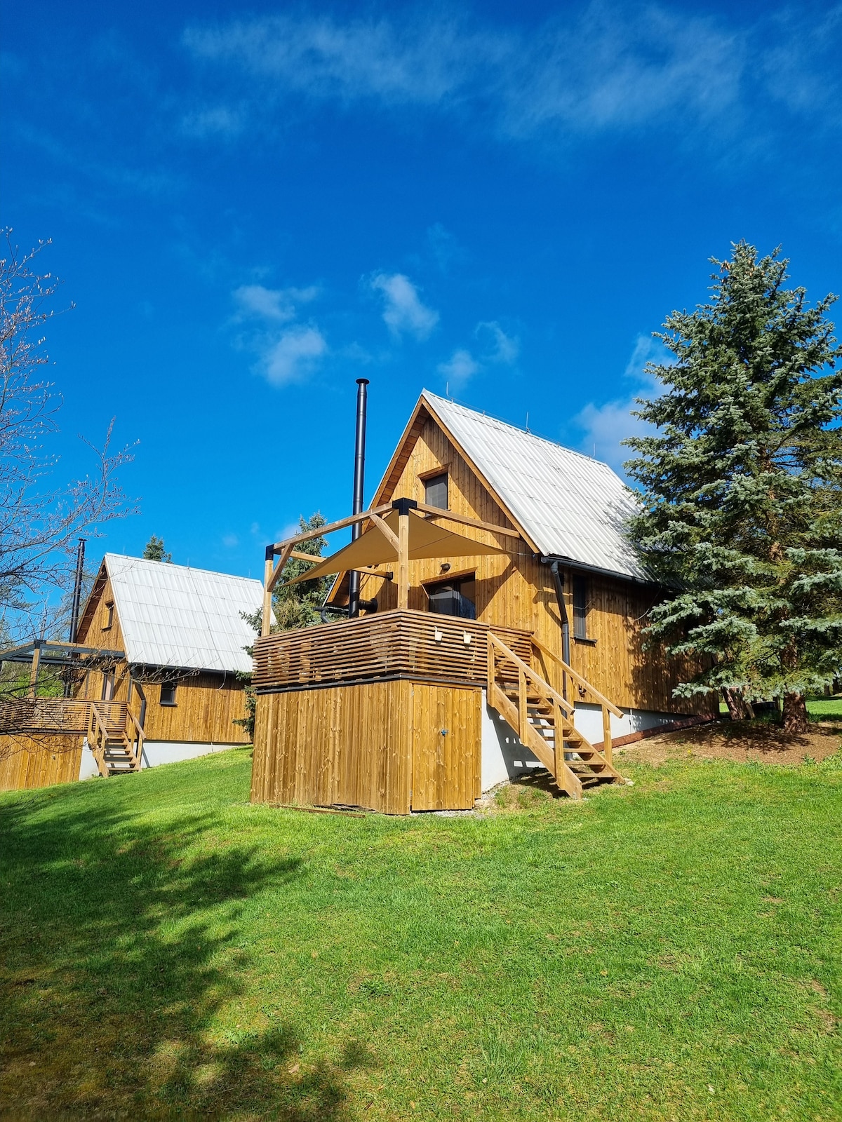 A two-story wooden cabin features a sloped roof and is surrounded by greenery. A wooden deck with steps leads to the entrance. Tall trees provide shade, and the sky is partly cloudy, contributing to a peaceful outdoor setting.