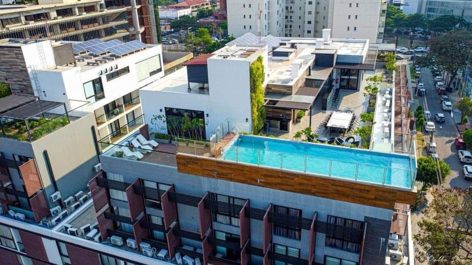 An aerial view captures a modern building complex with a rooftop infinity pool glistening under the sun. Surrounding greenery and sun loungers enhance the outdoor space. Nearby, various architectural styles create a vibrant urban landscape.