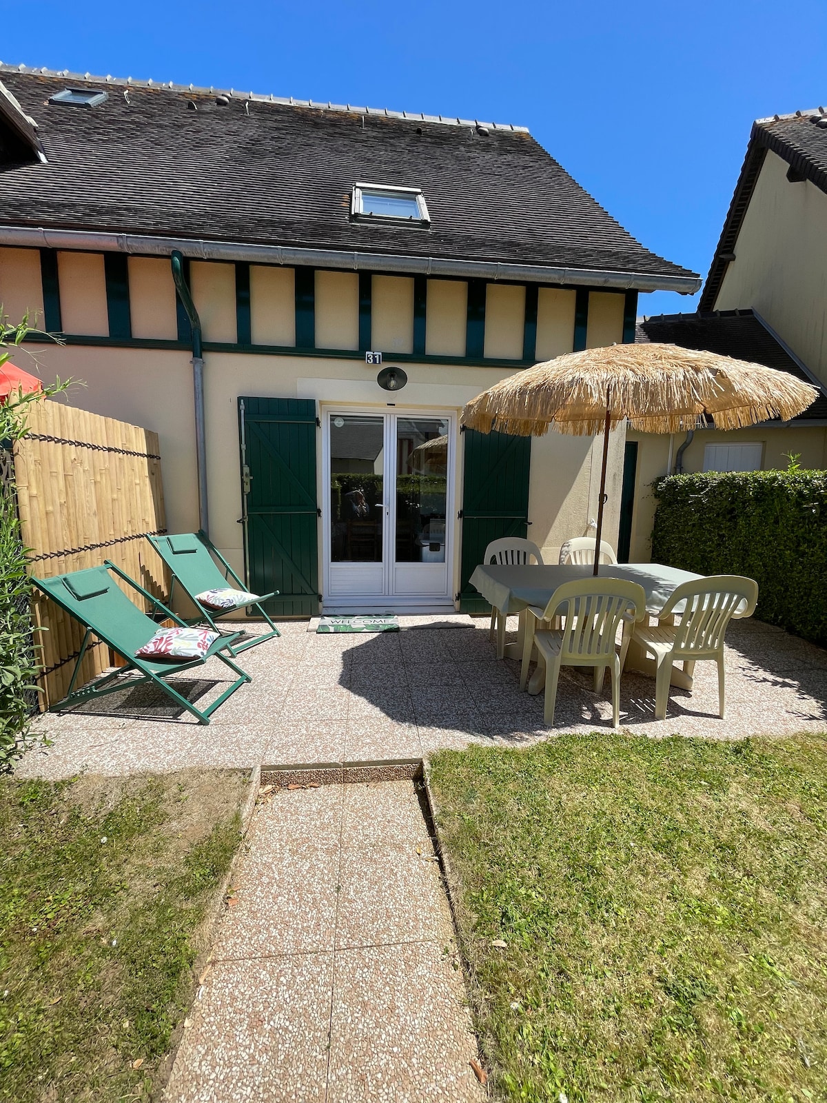 A private outdoor space is shown, featuring a table and chairs beneath a straw umbrella. Two reclining chairs are positioned nearby. The area is framed by greenery, offering a glimpse of the home's exterior with green shutters and a clear blue sky overhead.