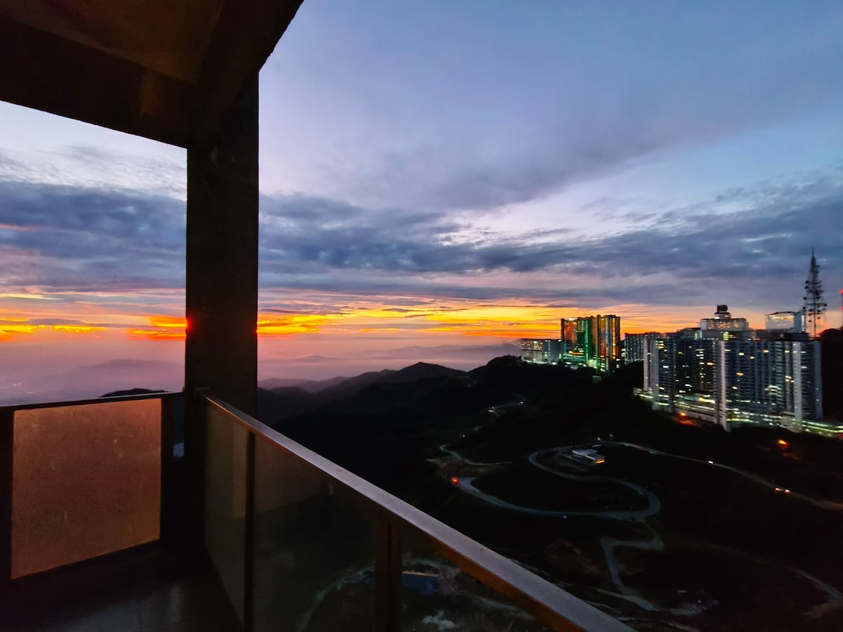 A stunning view from the balcony captures the vibrant hues of a mountain sunset, with shades of orange and purple painting the sky. Distant structures and hills create a serene backdrop, enhancing the expansive landscape visible beyond the glass railing.