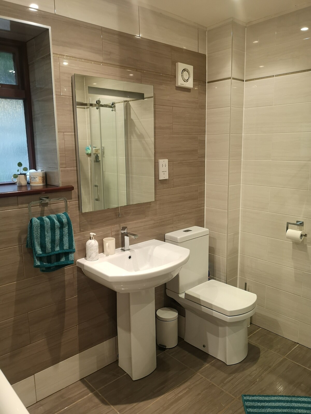 A modern family bathroom features a walk-in shower and a spacious bathtub. A large mirror reflects natural light, enhancing the neutral tile decor. Convenient toiletries are neatly arranged on a ledge, while a towel set hangs on a rack, providing easy access.