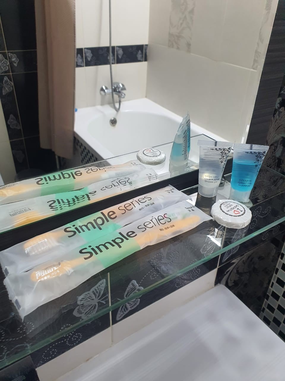 A neatly arranged bathroom counter is visible, with travel-sized toiletries placed on a glass shelf. A mirror reflects a bathtub and a shower fixture, while a packet of dental care items is positioned prominently. The black and white tile design adds a decorative touch.