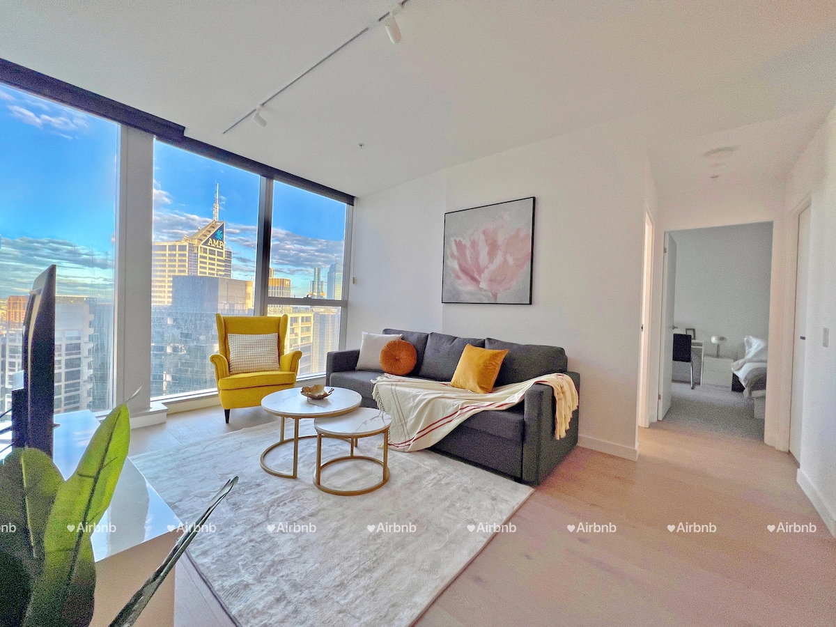 A spacious living room is illuminated by natural light from large floor-to-ceiling windows, offering views of the city skyline. A dark sofa is complemented by a vibrant yellow chair, while a round coffee table sits in the center, adorned with a decorative item.