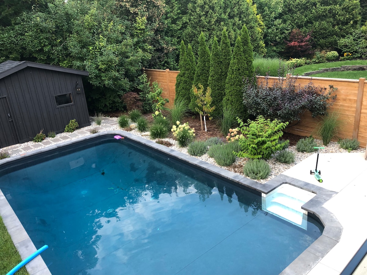 An inviting heated pool is surrounded by carefully landscaped greenery, featuring various plants and a tall wooden fence. A small shed is positioned to one side, and a few colored pool toys are lightly scattered in the water, adding a touch of playful color.