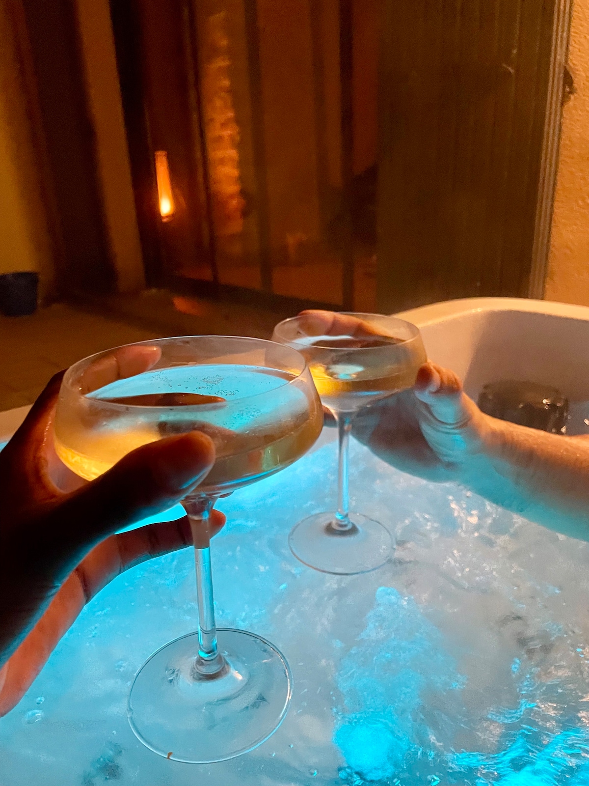 Two elegant glasses are raised in a toast above the bubbling surface of a jacuzzi, which is softly illuminated by blue lighting. The setting reflects a warm ambiance, with a faint outline of a wall and soft lighting in the background.