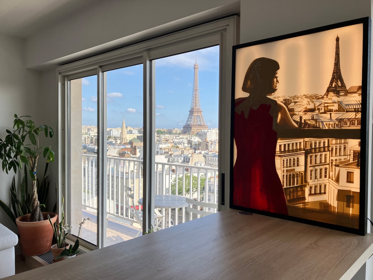A view of Paris from the apartment showcases the Eiffel Tower through large glass doors. A warm-lit art piece of a figure cast against an urban skyline adds depth. Potted plants and a wooden table complete the inviting interior, creating a serene atmosphere.