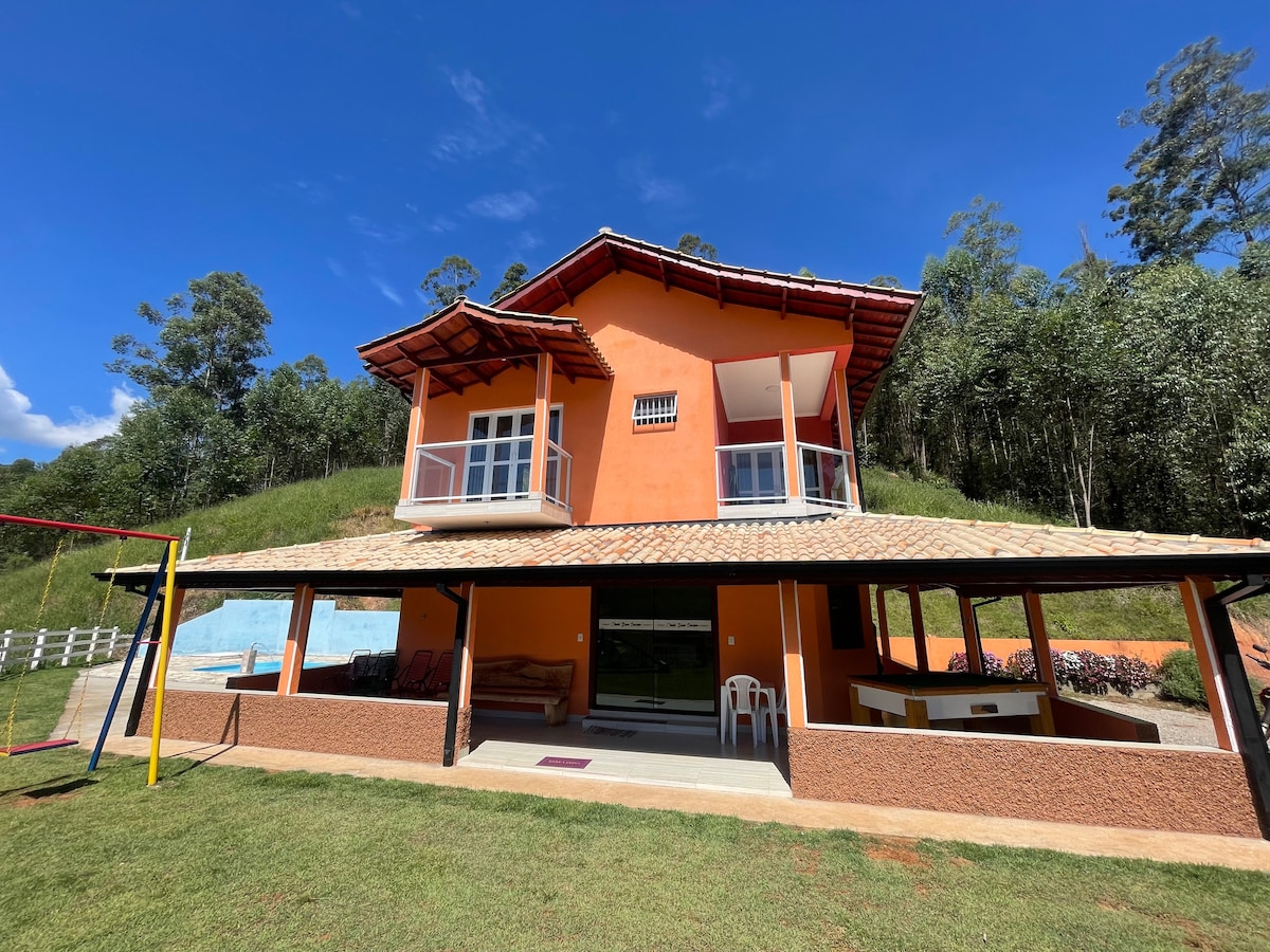 A two-story house features a vibrant orange exterior and a tiled roof. A covered patio extends from the house, providing a shaded seating area. Large windows and balconies invite natural light, while a well-maintained lawn surrounds the property, enhancing the outdoor space.