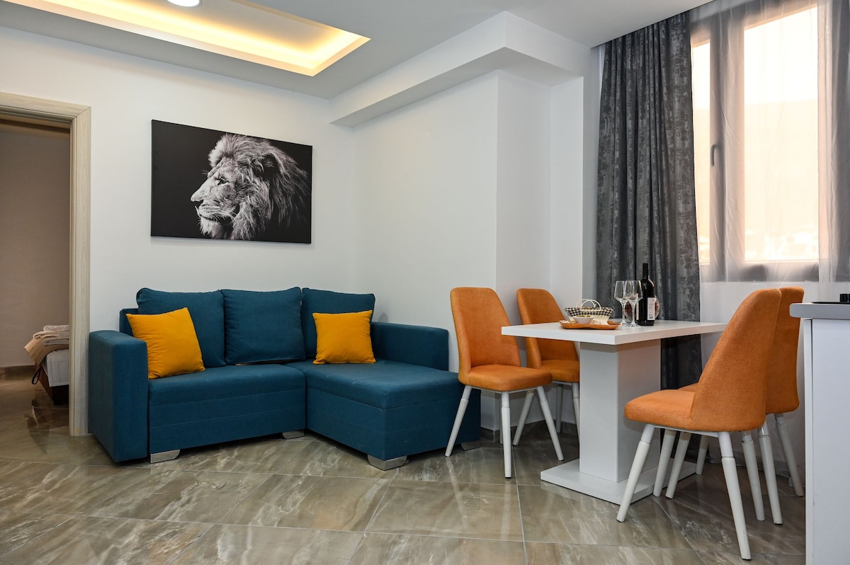 A modern living area is furnished with a teal sofa adorned with orange cushions. A white dining table surrounded by four orange chairs is positioned nearby. A striking black and white lion photograph decorates the wall, and natural light filters in through the window draped with gray curtains.