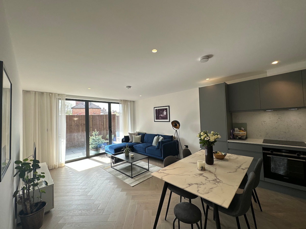 A modern living space features a blue sofa, a marble dining table, and sleek kitchen cabinetry. Large glass doors open to a private outdoor area, allowing natural light to fill the room. A decorative plant adds a touch of greenery to the contemporary design.