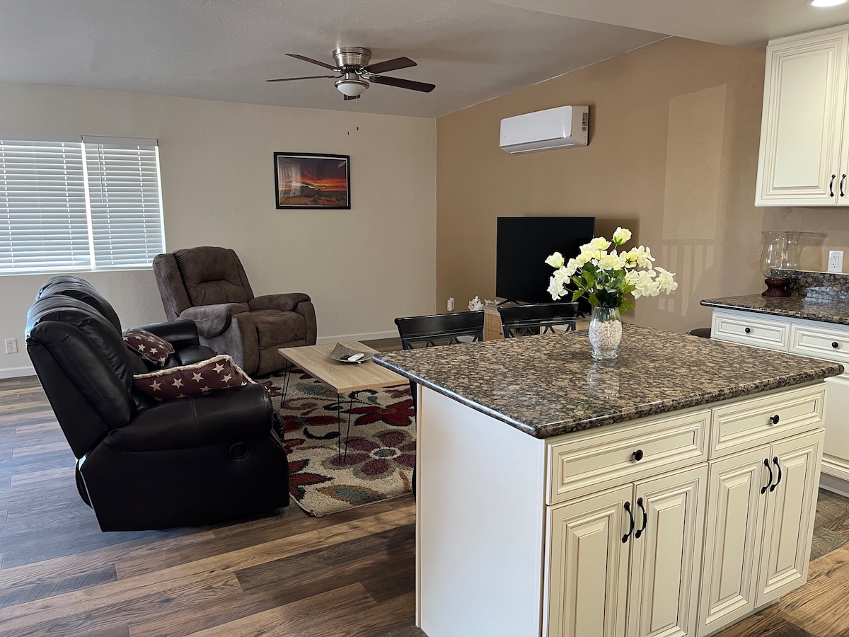 A modern living area features a comfortable recliner and a sofa, positioned around a coffee table. A flat-screen TV is mounted on the wall, with natural light coming through the window. A kitchen island with a granite countertop is visible in the foreground, adorned with a vase of flowers.