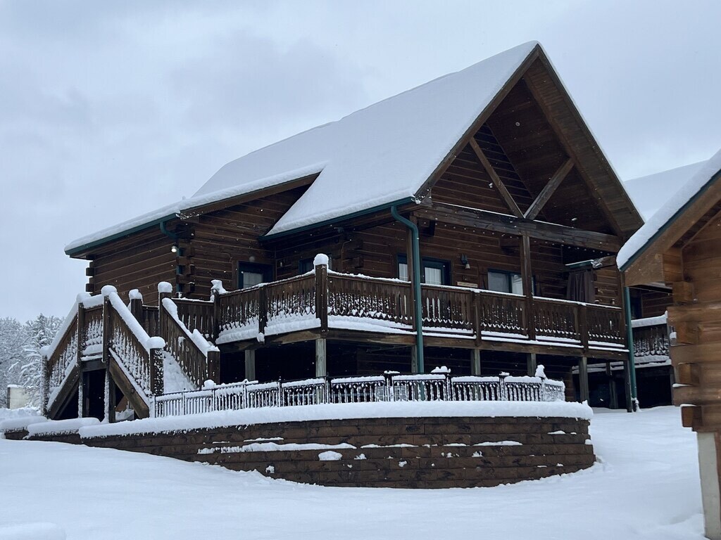 A spacious log cabin is surrounded by a blanket of fresh snow. The inviting front deck, bordered by rustic wooden railings, is accessible by a sturdy staircase. The steeply pitched roof adds character, while the winter landscape enhances the cabin's natural setting.