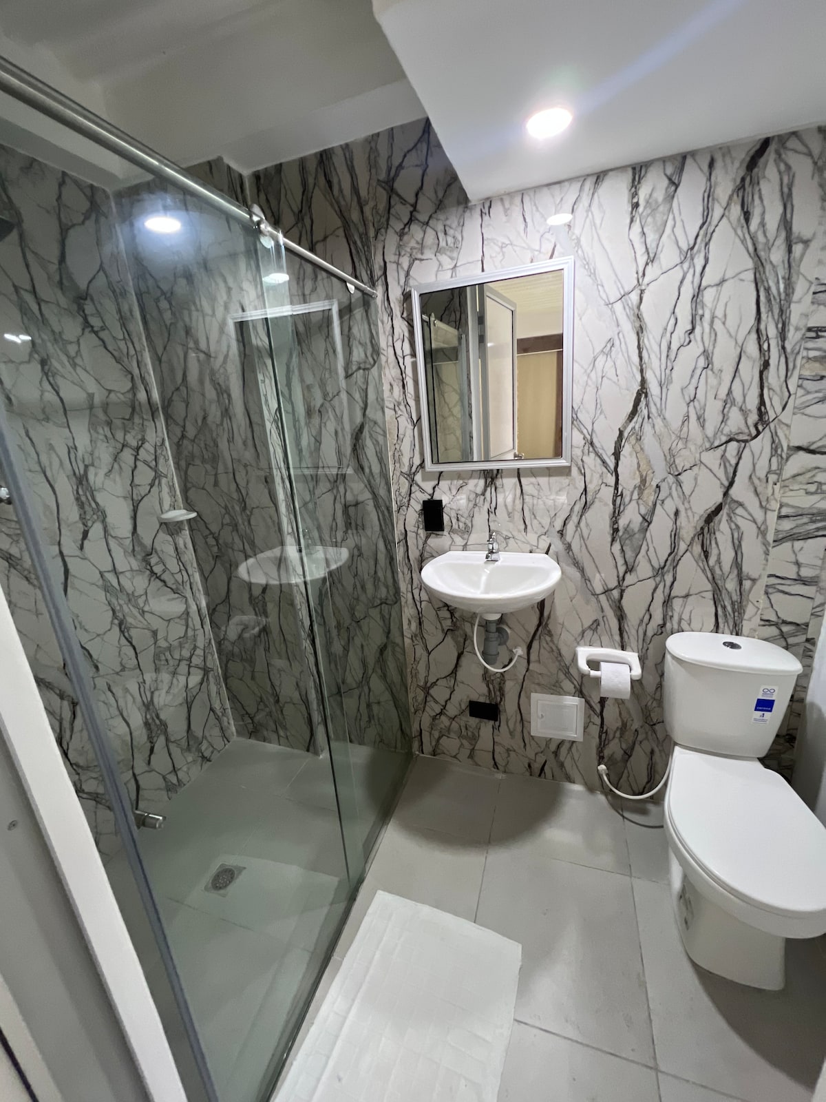 A modern bathroom features sleek marble-patterned walls and a glass shower enclosure. A small sink and mirror are mounted above a wall-mounted toilet. Soft lighting illuminates the space, enhancing its clean and contemporary design.