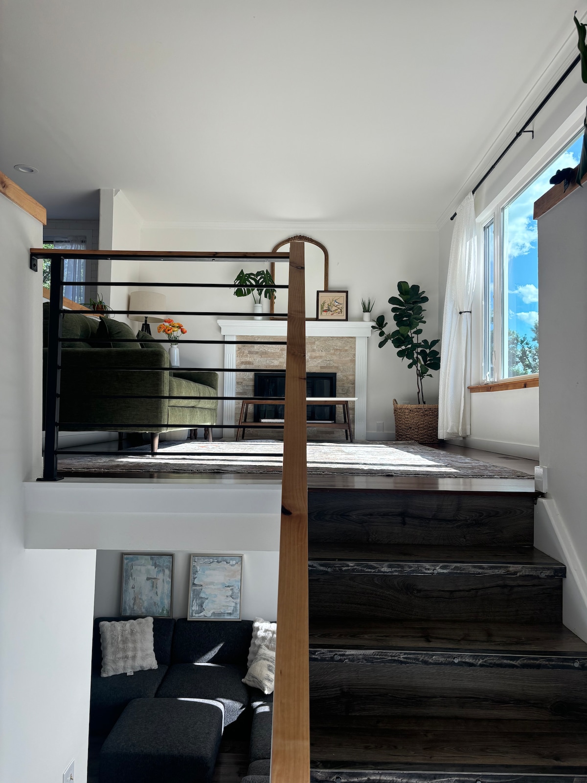 A staircase leads to a cozy living area with large windows, allowing ample natural light. Green plants adorn the space, complemented by a modern green sofa. A stone fireplace serves as a focal point, enhancing the clean and welcoming ambiance.