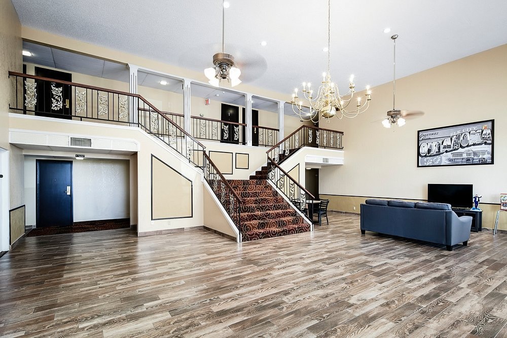 An open lobby area features polished wooden flooring and high ceilings. A grand staircase with decorative railings leads to an upper level. A seating area with a dark sofa and a cable TV is present, along with a dining space illuminated by a chandelier.