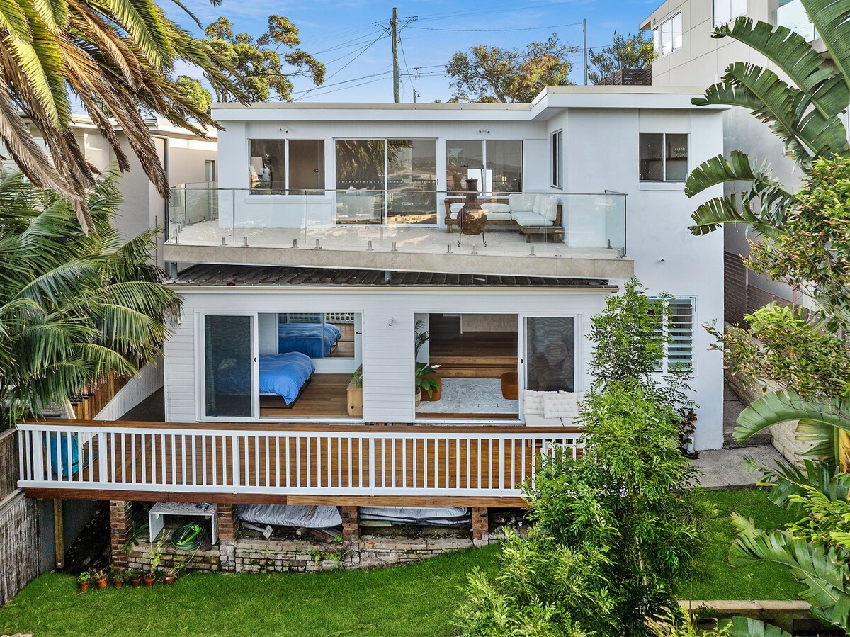 The exterior of a two-story apartment is showcased, featuring expansive glass doors that open onto a spacious back deck. Lush greenery surrounds the property, and various outdoor seating options are visible. The lower level includes a serene terrace garden, enhancing the inviting atmosphere.