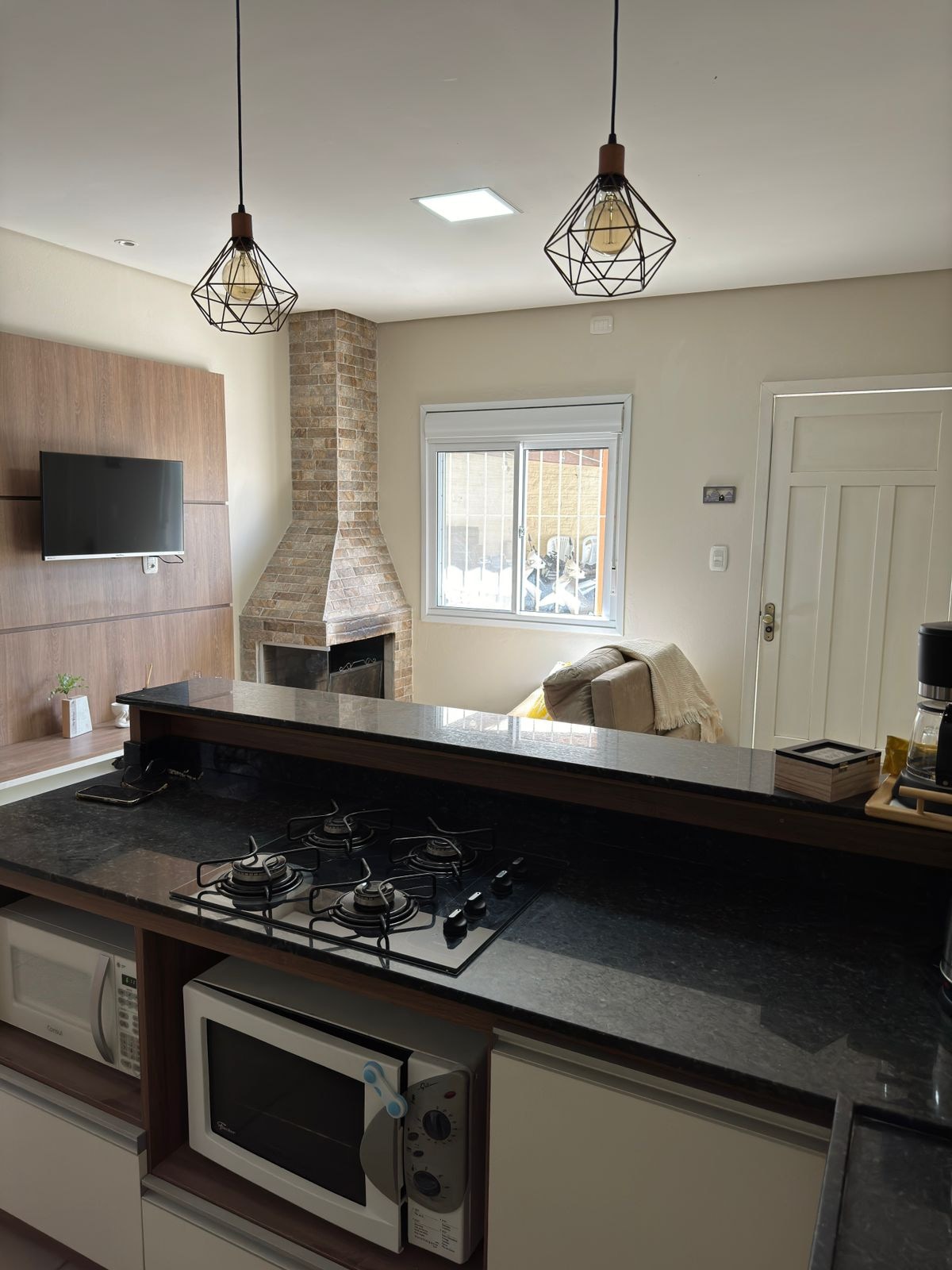 A modern kitchen layout is showcased, featuring a dark countertop with a gas stove and a microwave. A cozy living area is visible, highlighted by a brick fireplace and a mounted TV. Natural light enters through a window with a simple curtain.