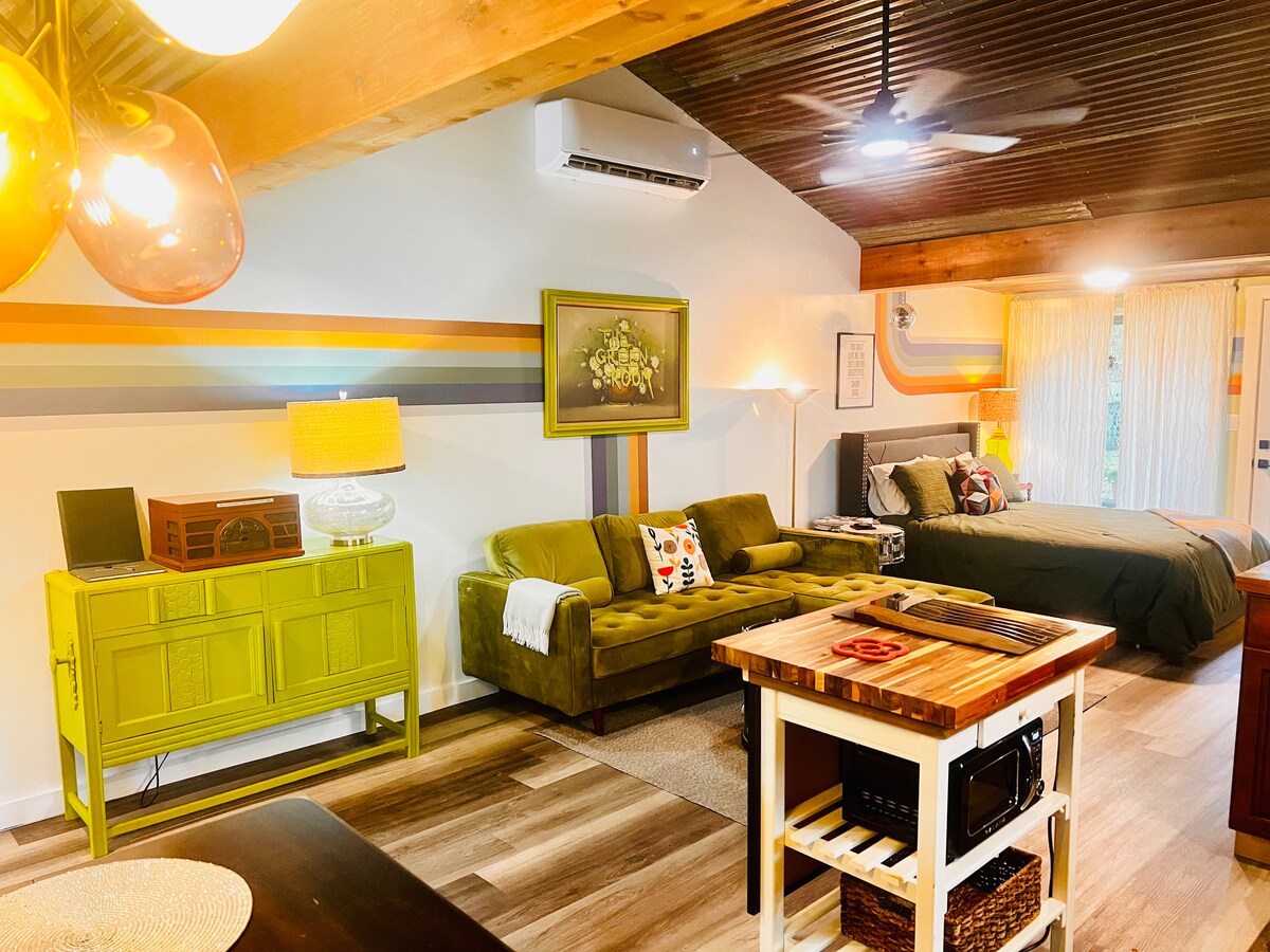 A vibrant living space features a green velvet sofa placed next to a bright green cabinet. A vintage record player is visible on the cabinet. Behind, a spacious bed is complemented by soft linens, with ample natural light coming through sheer curtains.