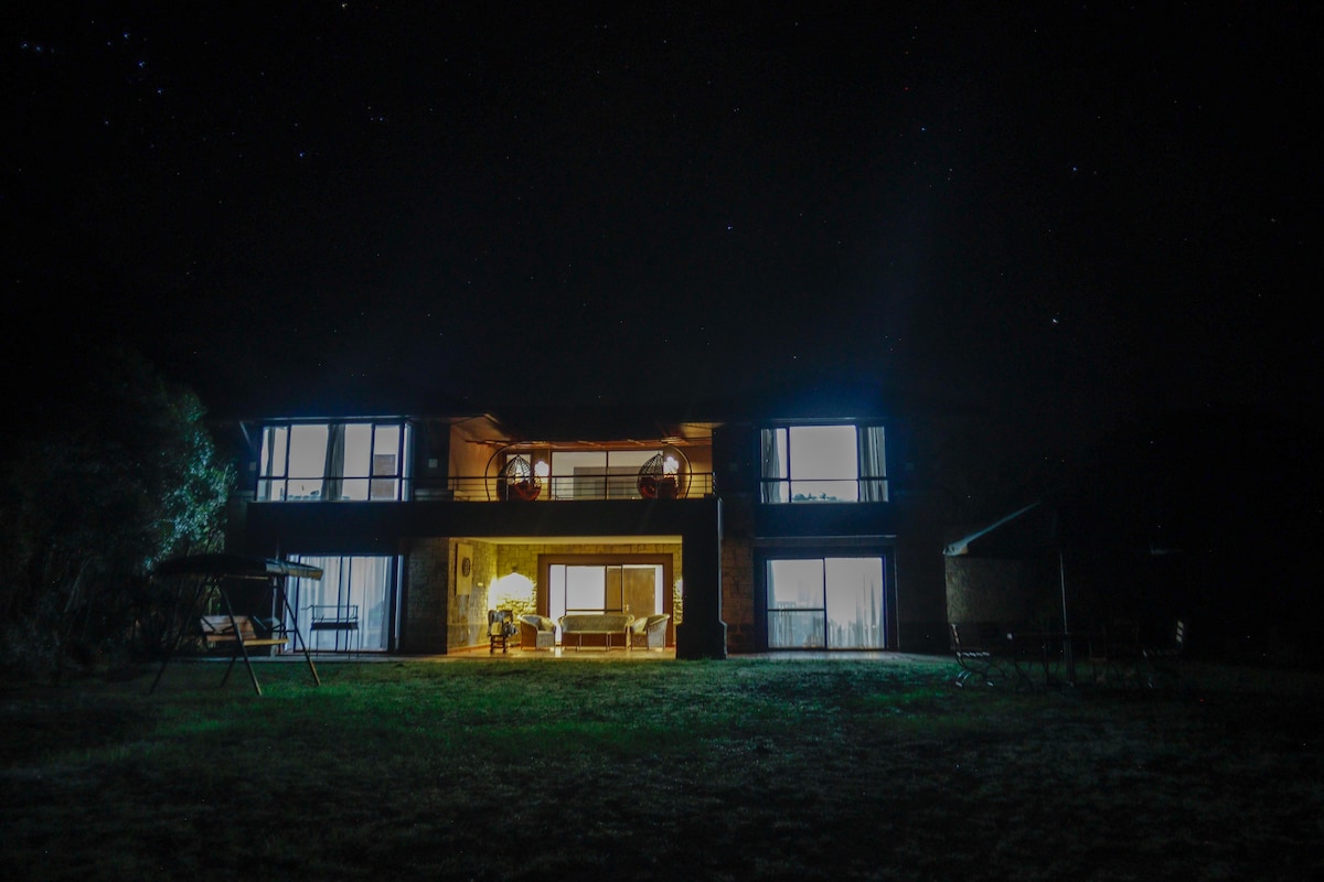 The villa is shown from the exterior at night, illuminated by soft light from the windows. A spacious terrace is visible, flanked by trees, and the starry sky adds a serene backdrop to the scene.