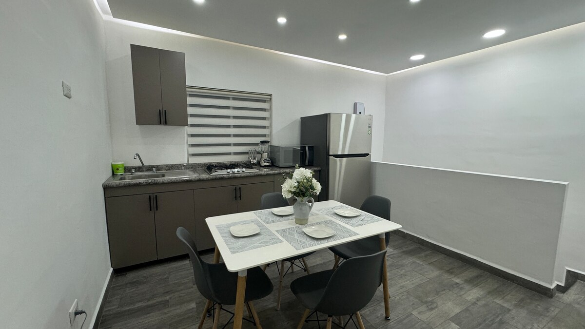 A modern kitchen area is visible, featuring a dining table with four chairs. The countertops are equipped with appliances, including a refrigerator. Soft overhead lighting enhances the neutral color scheme, while a bouquet of flowers adds a touch of decor to the table.