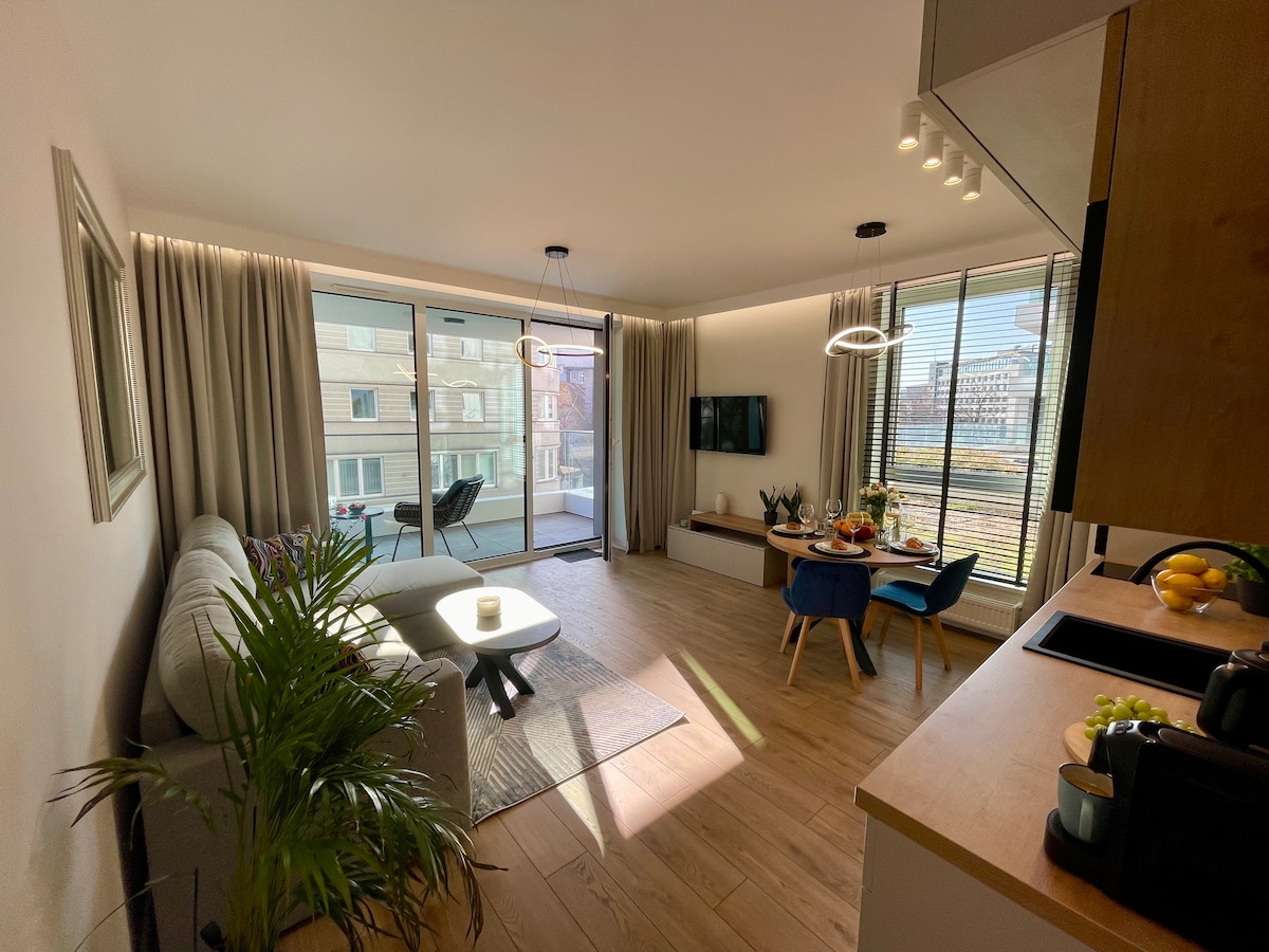 A bright living space features a comfortable sofa and a small dining area with blue chairs. Large windows allow natural light to fill the room, revealing a view of the exterior. A decorative rug and potted plant add subtle touches to the welcoming atmosphere.