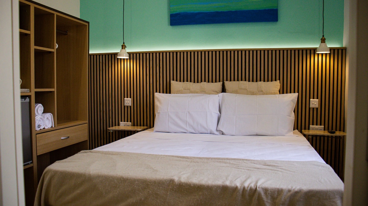 A cozy bedroom is showcased with a spacious bed featuring crisp white linens. Soft lighting is provided by hanging bedside lamps. A wood-paneled headboard adds texture, while a simple piece of wall art in soothing colors decorates the space. Stacked towels are neatly arranged on a nearby shelf.