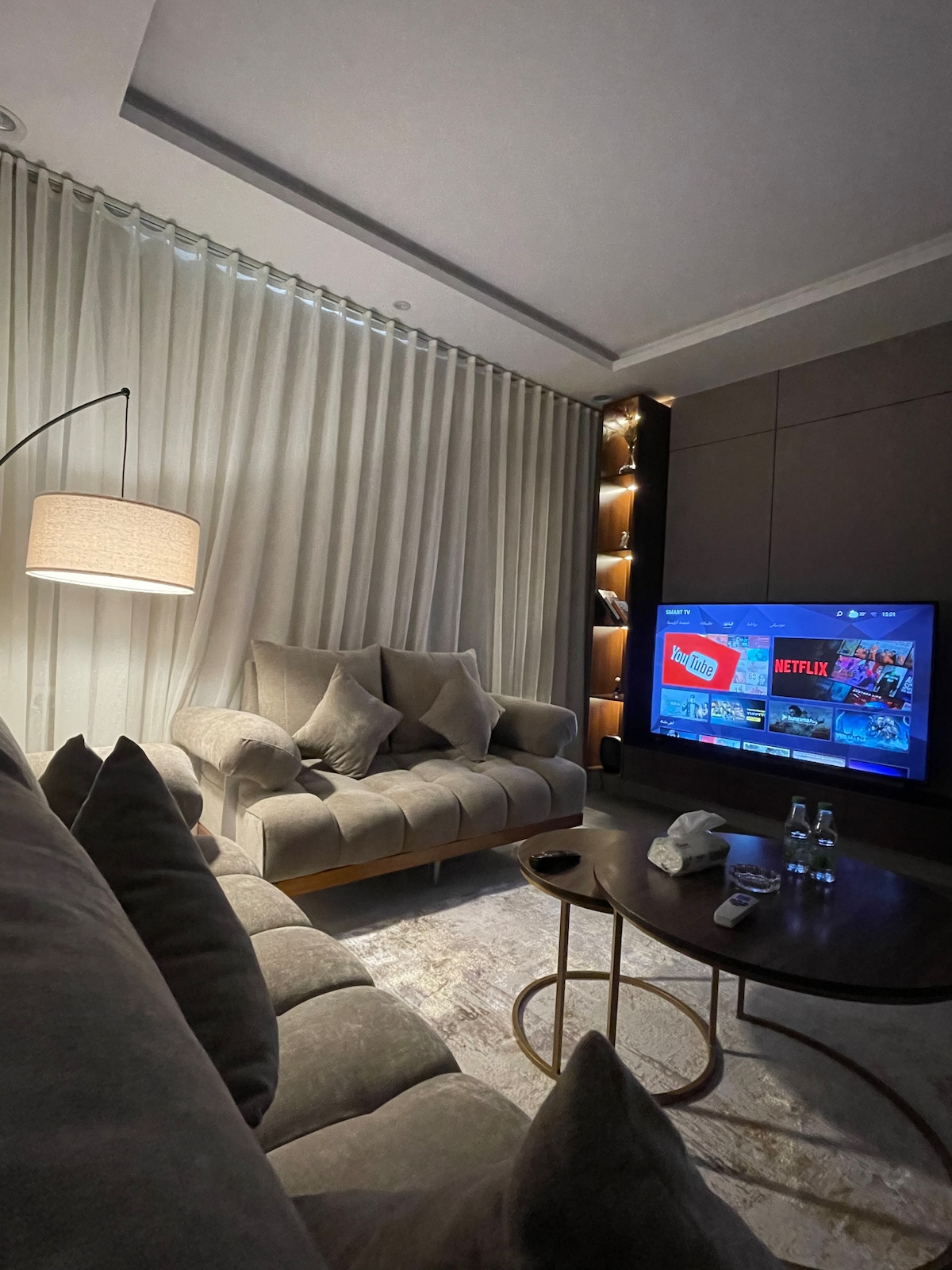 A cozy living area features a plush sofa with multiple cushions, complemented by a stylish floor lamp. A sleek coffee table holds two bottles of water. The wall-mounted television displays a streaming service, surrounded by modern decor and soft ambient lighting from the built-in shelves.