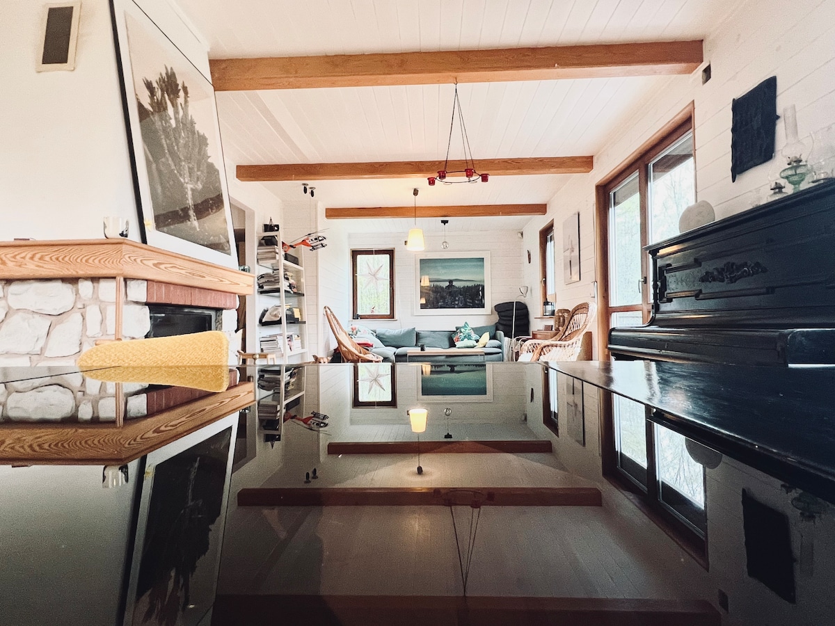 A spacious living area is depicted with wooden beams extending across the ceiling. A reflective surface showcases the room’s light fixtures. The fireplace adds warmth, while comfortable seating and decor create a relaxed atmosphere. Large windows invite natural light into the space.