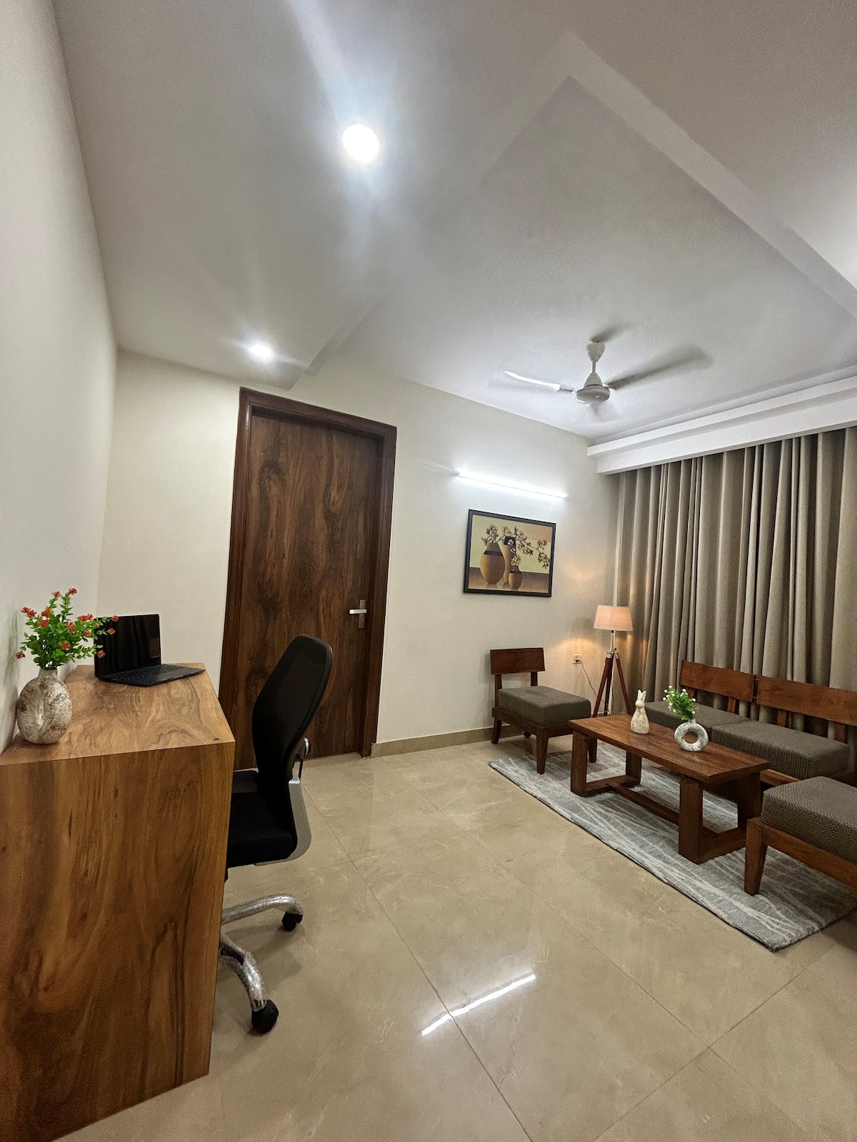A well-lit living space features a wooden desk and chair, complemented by two wooden benches and a coffee table. A decorative plant adds a touch of greenery. Soft lighting from a nearby lamp creates a comfortable ambiance, while elegant curtains frame a large window.