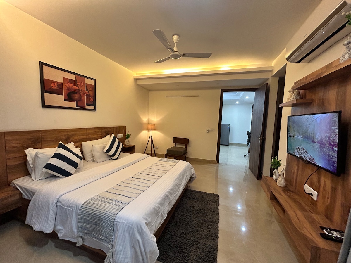 A spacious sleeping area is furnished with a large bed, featuring white linens and contrasting striped pillows. A soft rug lies on the polished floor, and artwork adorns the wall above the bed. A flat-screen TV is mounted nearby, and a ceiling fan provides comfort.