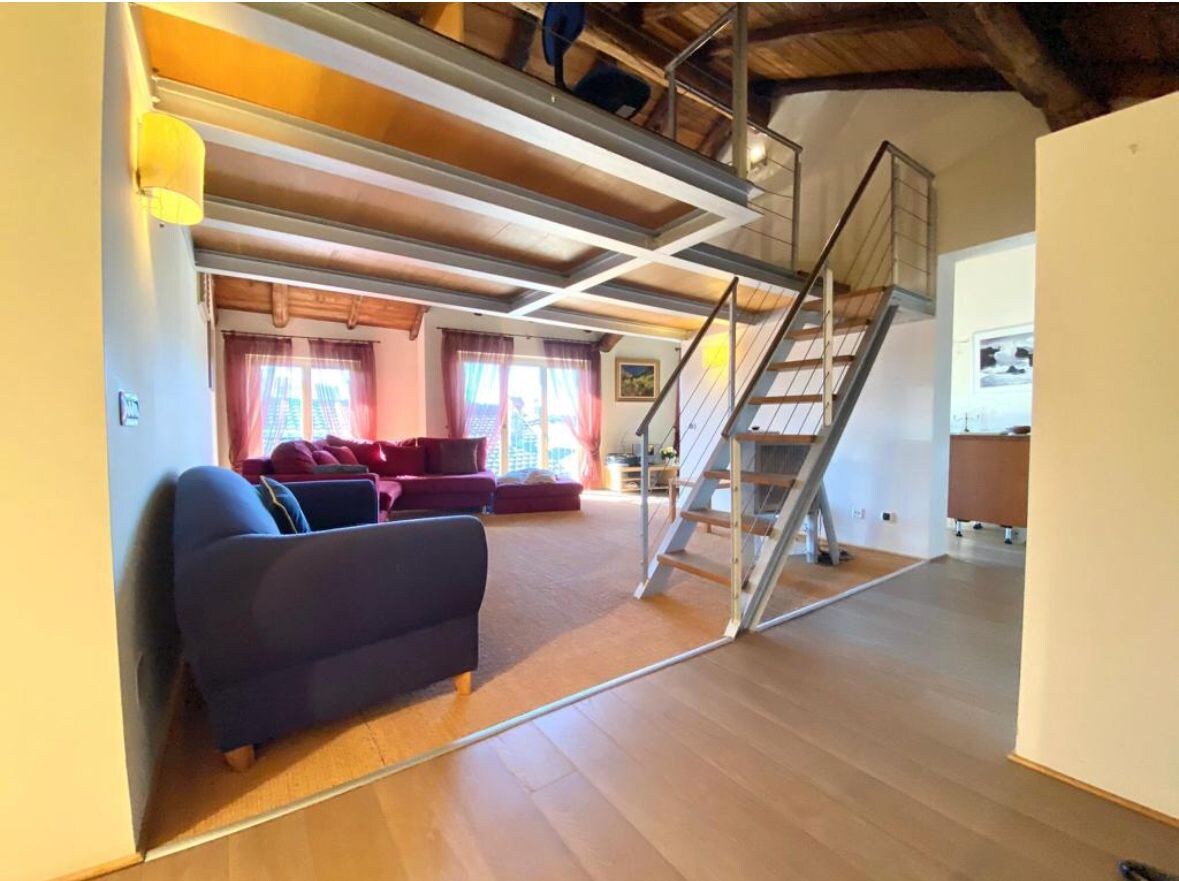 A spacious living area is presented with a cozy arrangement of seating, including a dark blue sofa and a red sofa. Tall windows allow natural light to fill the room, which is complemented by warm wood beams overhead. A staircase leads to an upper level, enhancing the overall openness.