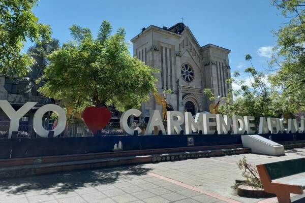 A large sign displaying 'YO ♥ CARMEN DE APICALA' is prominently featured in front of a historic church. The church's architectural details are highlighted against a clear blue sky and surrounded by lush green trees.