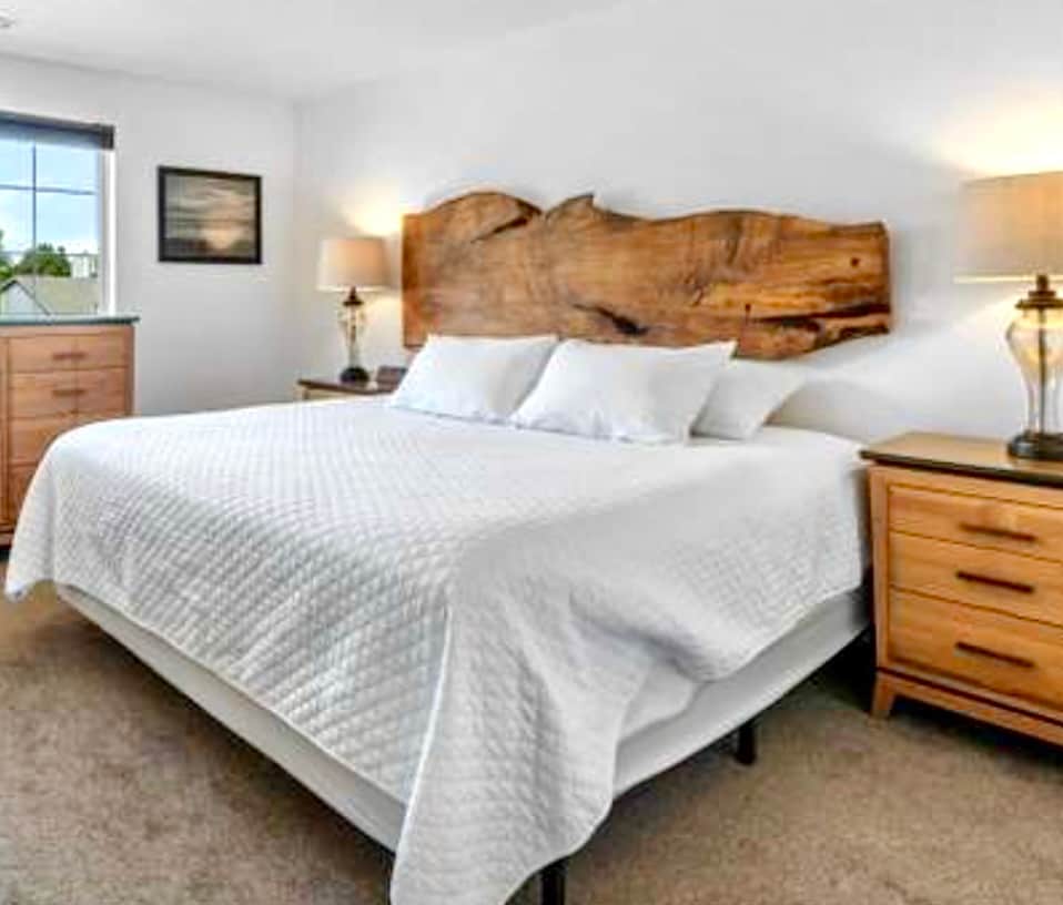 A cozy bedroom features a spacious bed with a quilted white coverlet, complemented by natural wood accents on the headboard. Two bedside lamps illuminate the space, and a window allows natural light to brighten the room. A dresser is positioned nearby.