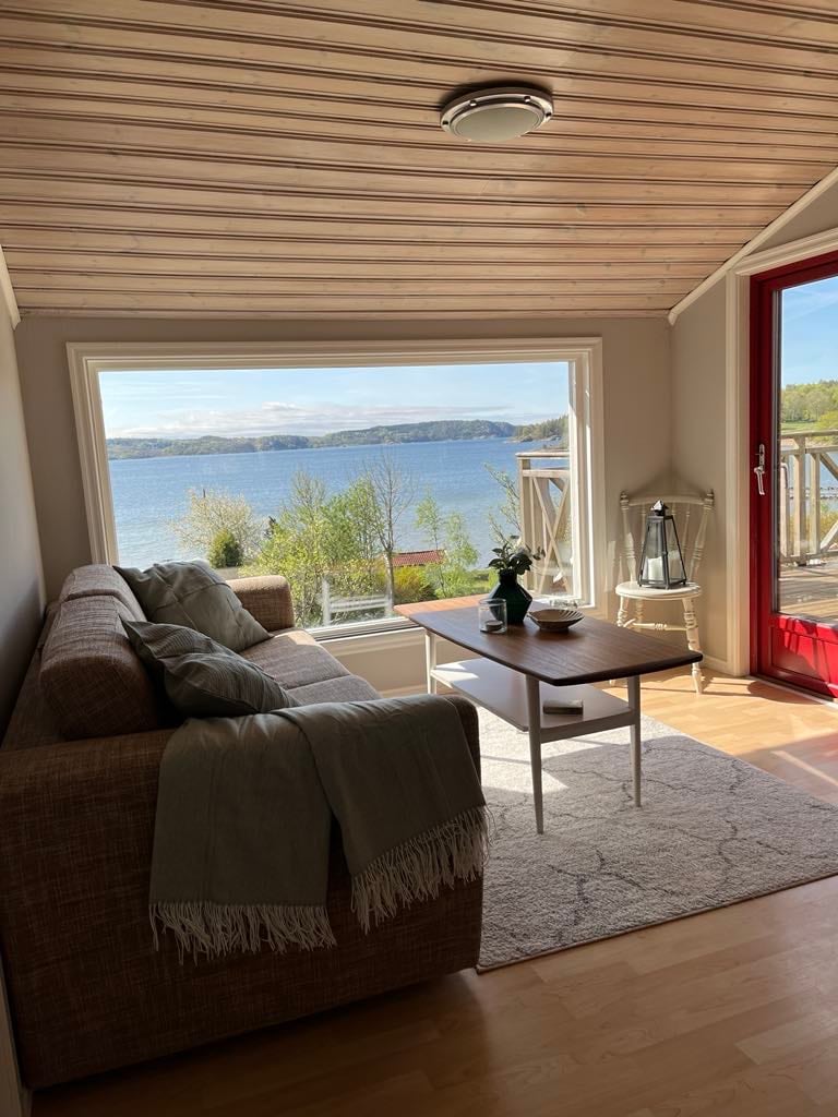 A cozy sitting area features a comfortable sofa adorned with a soft throw. A table holds a small plant, while large windows provide a view of the serene sea and surrounding greenery. Natural light illuminates the warm wooden floors.