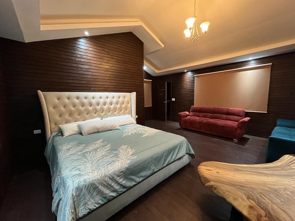 A spacious sleeping area features a large upholstered bed with a light blue quilt adorned with a floral pattern. The rich wooden walls provide warmth, complemented by a sofa in soft red and a unique wooden table. Soft lighting enhances the relaxing ambiance.