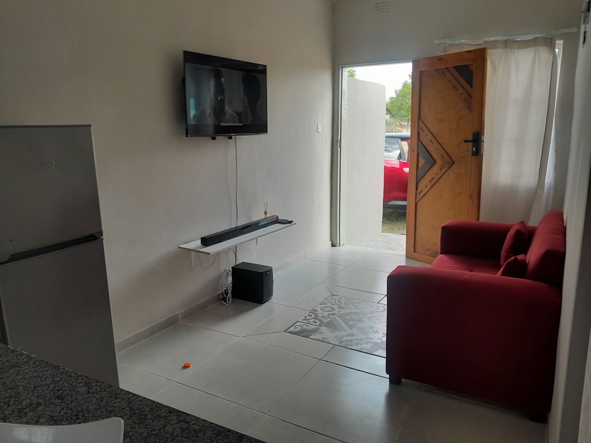 A living area features a red couch positioned next to a wall-mounted television. A sleek fridge is visible in the kitchen area, and natural light enters through a partially open door. The floor displays a patterned tile, adding character to the space.
