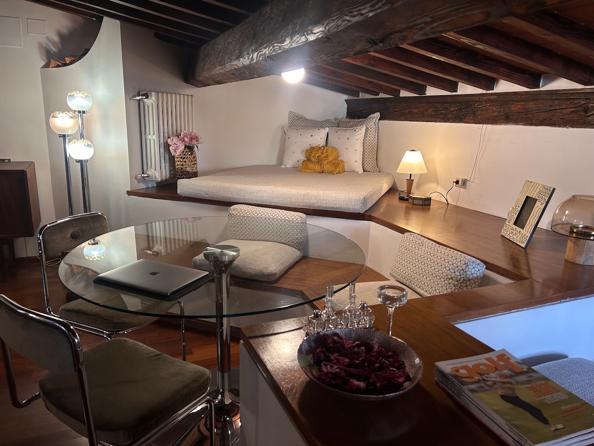 An interior view showcases a loft space with a raised sleeping area featuring a spacious mattress adorned with decorative pillows. A circular glass dining table surrounded by stylish chairs is positioned near a modern light fixture, while warm wooden beams add character to the ceiling.