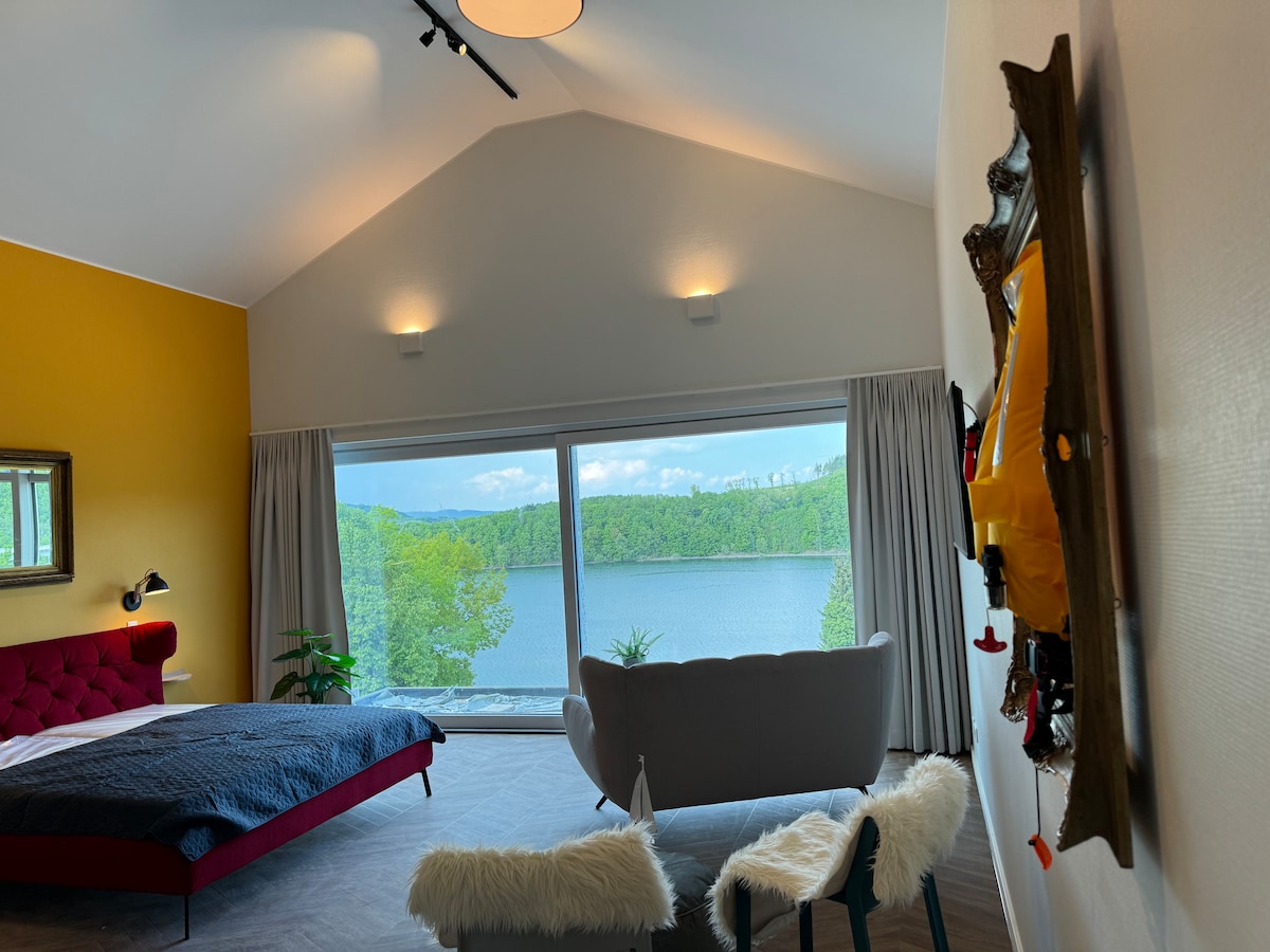 A spacious bedroom features large floor-to-ceiling windows showcasing a serene view of the lake and surrounding greenery. A comfortable red bed and a stylish gray armchair are positioned to take advantage of the natural light, complemented by warm yellow accent walls.