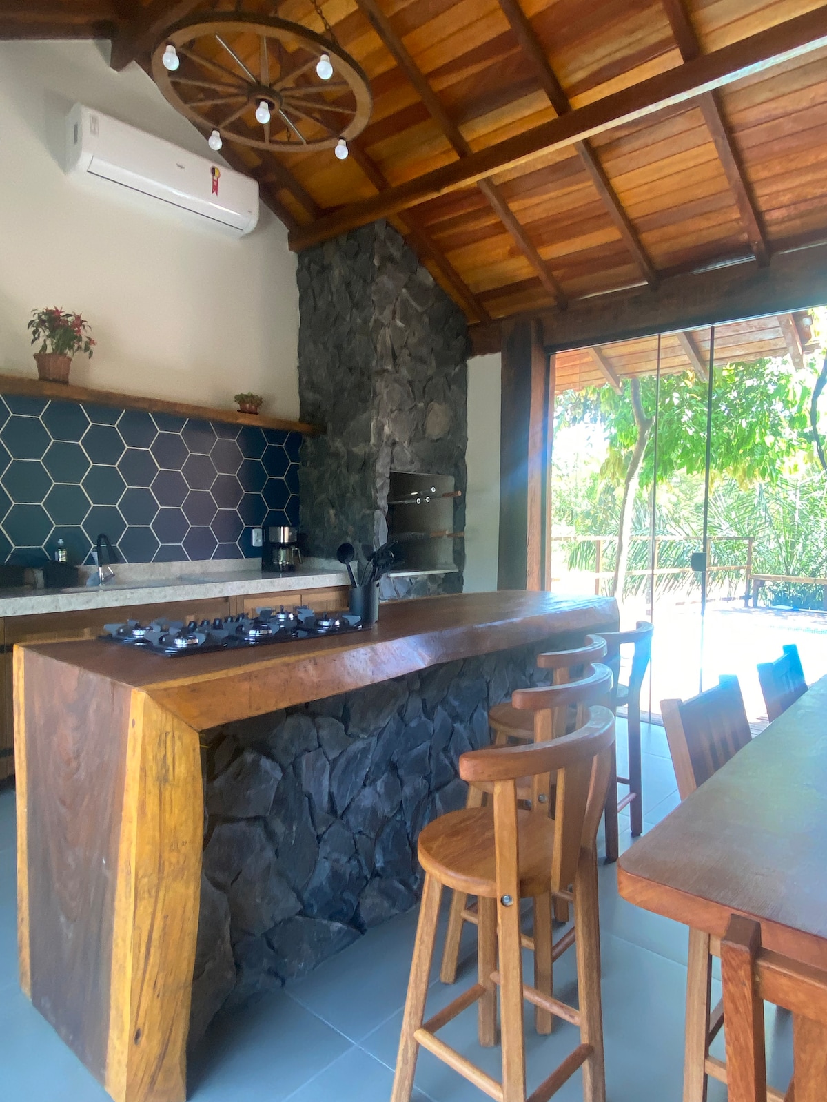 A modern kitchen is featured with a wooden, stone-accented island bar, accompanied by tall wooden stools. A hexagonal tile backsplash adds texture, while large glass doors enhance natural light and provide a view of the outdoor area surrounded by greenery.