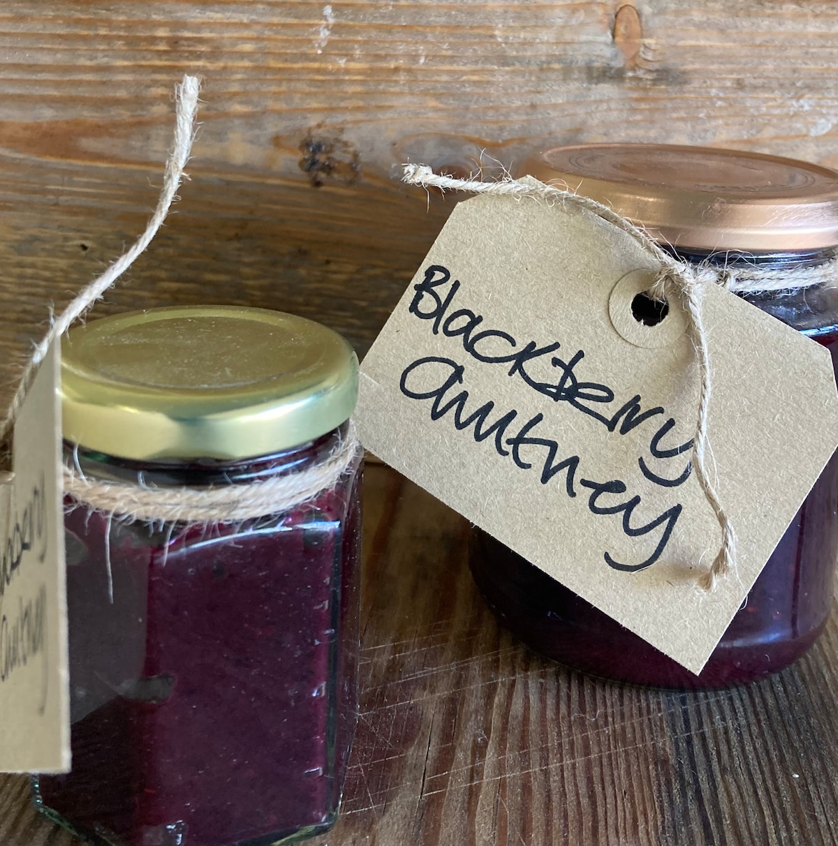 Two glass jars of homemade blackberry jam are displayed against a rustic wooden background. Each jar is topped with a golden lid and is wrapped with twine, featuring labels that read 'Blackberry' and 'Amber'. The deep purple color of the jam is easily visible.